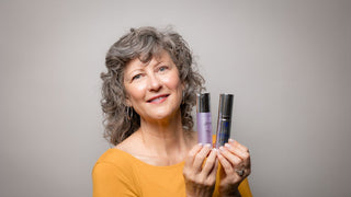 Woman in mustard shirt holding two skincare products with grey background