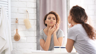 Woman looking in mirror inspecting skin and touching cheek with bathroom products on the wall