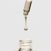 Close-up of a serum dropper releasing a golden oil drop above an open glass bottle on a light gray background