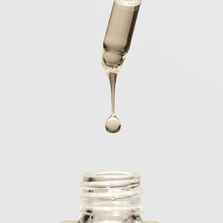 Close-up of a serum dropper releasing a golden oil drop above an open glass bottle on a light gray background