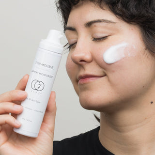Woman with eyes closed holding CO2Lift Skin Mousse Carboxy Moisturizer canister beside her face with foam product applied to cheek, expressing relaxation during anti-aging skincare routine