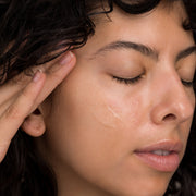 Close-up of a woman with curly dark hair and eyes closed applying Hale & Hush Quiet Wash cleanser to her cheek, showing the product's lightweight clear gel texture on skin