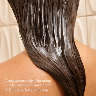 Back view of a person with long dark hair applying SEEN Conditioner in the shower, with white conditioner visible throughout wet hair and the instruction to leave on for 2–3 minutes before rinsing
