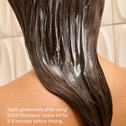 Back view of a person with long dark hair applying SEEN Conditioner in the shower, with white conditioner visible throughout wet hair and the instruction to apply generously after using SEEN Shampoo and leave on for 2–3 minutes before rinsing.