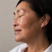 Side profile of woman with dark hair and glowing even skin tone with eyes closed, showing Dr.esthe Anti-Mela Toning Serum brightening and hyperpigmentation results on neutral background