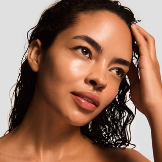 Woman with glowing hydrated skin and wet curly hair touching her face against a light gray background