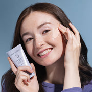 Woman applying Face Reality PowerMineral SPF 50 acne-safe mineral sunscreen to cheek while holding tube against blue background