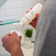 Hands dispensing Face Reality Glycolic-Lactic Toner onto a cotton pad in bathroom – AHA exfoliating toner for skin renewal