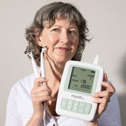 A woman with short gray curly hair holding the MyoLift TW device displaying Rejuv Level 2, Intensity 176, and Time 20:00, with the microcurrent probes resting against her neck, against a light gray background.