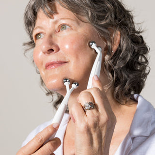 A woman with short gray curly hair applying the MyoLift TW microcurrent probes to her cheek and jaw, holding one probe in each hand against a light gray background.