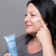 Woman applying GlyMed Plus Anti-Aging Exfoliant Masque to cheek while holding the light blue tube