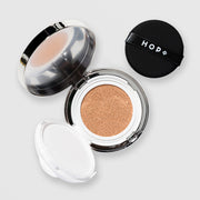 HOP+ Volume Shield Cushion open showing beige foundation-soaked cushion sponge, removable inner tray, black branded puff and mirrored lid, silver compact