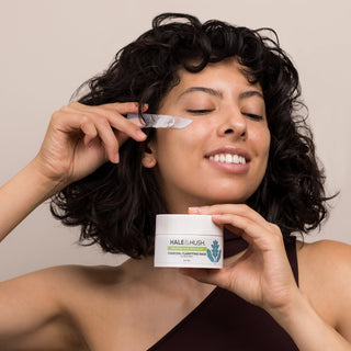 Woman with curly dark hair smiling while applying Hale & Hush Charcoal Clarifying Mask to her cheek with a spatula, holding the white 3oz jar