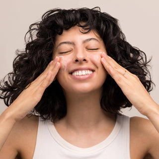Smiling woman with curly dark hair massaging Hale & Hush Clarify Wash facial cleanser onto both cheeks with eyes closed, neutral background