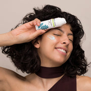 Smiling woman with curly dark hair holding Hale & Hush Hush Hydrate Gel Moisturizer tube to her forehead with a cream swatch applied to her cheek