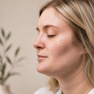 Side profile of blonde woman with eyes closed and a drop of Hale & Hush Rare C Serum applied to cheek, showing lightweight clear formula on sensitive skin