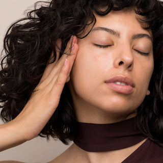 Woman with curly dark hair and glowing skin gently pressing hand to cheek with eyes closed, showing Hale & Hush Soothe Essence Serum absorption