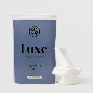 Art of Skin Care Luxe Toner Pads 100-count in a slate blue resealable pouch with AS monogram logo, displayed alongside a stack of white textured toner pads, against a light gray background.