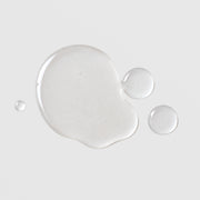 Michele Corley Calming Cleansing Oil and Makeup Remover texture showing lightweight clear oil droplets on grey background.