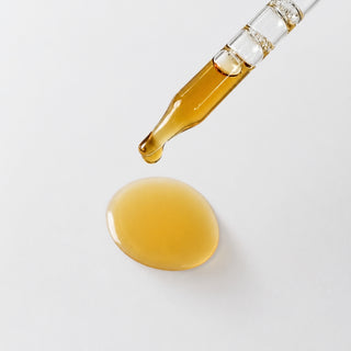 Michele Corley Licorice Root Additive texture showing golden amber serum drop from glass dropper on grey background.