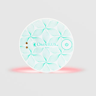 Omnilux Mini Blemish Eraser LED device in a round shape with a white and teal floral line pattern, power button, and visible red LED light glow along the bottom edge, displayed against a light gray background.