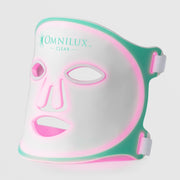 Omnilux Clear LED face mask in white silicone with a teal border and teal logo, glowing pink and red from within through the eye, nose, and mouth openings, with white elastic head straps, displayed against a light gray background.