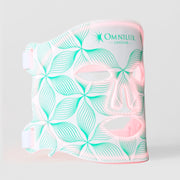 Omnilux Contour Face LED mask in white silicone with a teal floral line pattern, glowing pink through the eye, nose, and mouth openings, with white elastic head straps, displayed against a light gray background.