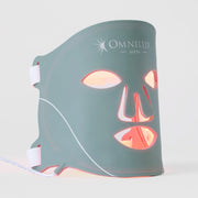 Omnilux Men LED face mask in slate teal silicone with white logo, glowing red and amber through the eye, nose, and mouth openings, with white elastic head straps, displayed against a light gray background.