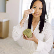 Woman in a white robe applying a Sorella Apothecary peel pad to her face while holding the jar in a bright bathroom