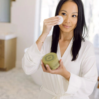 Woman in a white robe applying a Sorella Apothecary peel pad to her face while holding the jar in a bright bathroom