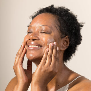 Smiling woman with natural curly hair applying iS clinical brightening complex cream to her glowing face with both hands against a neutral beige background