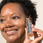Smiling woman with natural hair applying iS Clinical Youth Eye Complex under her eye while holding the bottle