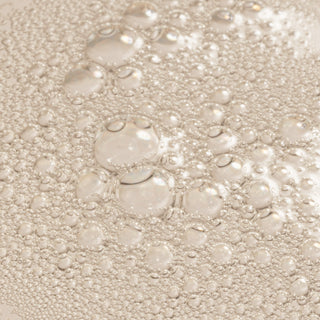 Close-up of Rhonda Allison Mandelic Defense Tonic liquid texture showing clear bubbling toner formula on a warm beige surface
