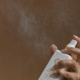 A person spraying Sorella Apothecary Watermelon Mint Hydrating Mist onto skin, with a fine mist visible in the air against a warm tan background.