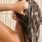 Back view of a person lathering SEEN Shampoo into long dark wet hair in the shower, with visible suds throughout, and the instruction to apply to wet hair, lather, and rinse.