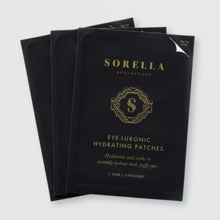 Three packets of Sorella Apothecary Eye-Luronic Hydrating Patches in black and gold foil packaging on a light gray background