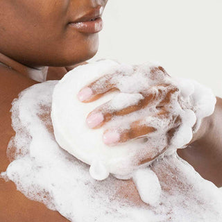 Close-up of a person lathering rich white foam from a loofah onto their shoulder, demonstrating the Farmhouse Fresh Sunshine Silk Soothing Body Wash in use.
