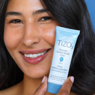 
Woman with glowing skin holding TiZO3 Primer Sunscreen Tinted SPF 40 facial mineral sunscreen tube against blue background