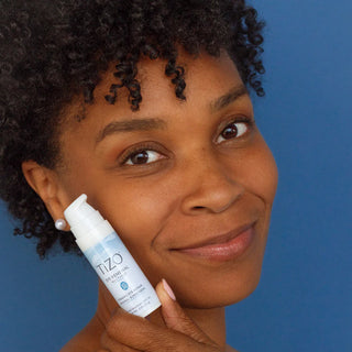 Woman with glowing skin holding TiZO Eye Renewal Non-Tinted SPF 20 firming eye repair mineral sunscreen against blue background