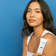 Woman with radiant skin holding TiZO Photoceutical AM Replenish Lightly Tinted SPF 40 moisturizing mineral sunscreen against blue background