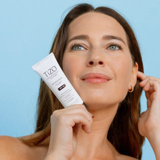 Woman with glowing skin holding TiZO Photoceutical AM Replenish Non-Tinted SPF 40 moisturizing mineral sunscreen against blue background
