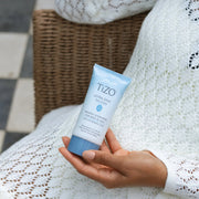 Hand holding TiZO Ultra Zinc Non-Tinted SPF 40 mineral sunscreen tube for body and face while seated in wicker chair with white knit blanket