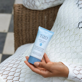 Hand holding TiZO Ultra Zinc Non-Tinted SPF 40 mineral sunscreen tube for body and face while seated in wicker chair with white knit blanket