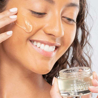 A smiling woman applying a streak of Farmhouse Fresh Watercress Hydration Cascade Geléé to her cheek while holding the open jar, against a white background