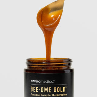 Close-up of golden raw Manuka honey dripping from a spoon into an open Enviromedica Bee-Ome Gold jar, showcasing rich texture of functional honey for the microbiome