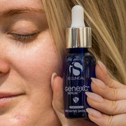 Woman with glowing skin holding iS Clinical GeneXC Serum cobalt blue dropper bottle against her cheek in natural light