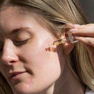 Woman in white shirt applying iS Clinical C Eye Serum Advance+ dropper to her cheekbone area in natural sunlight