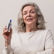 Smiling woman with silver hair holding iS Clinical Youth Serum bottle while seated against a neutral beige background