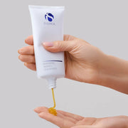 Hand holding a white tube of  iS Clinical Warming Honey Cleanser being squeezed onto the palm against a light gray background.