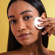 Le Mieux Skin Clarifying Pad in use showing woman applying round white exfoliating pad to cheek against warm yellow background. Available at Art of Skin Care
